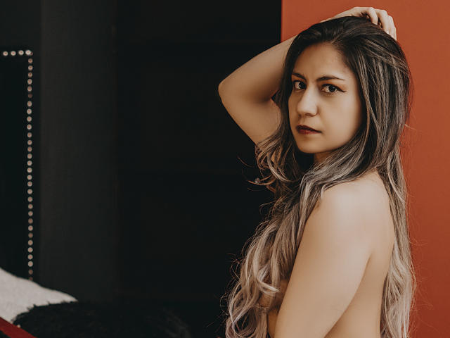ValeriaLoren, avec de longs cheveux ondulés, gris à blonds, nue, regardant par-dessus son épaule, une main dans les cheveux, yeux marrons, léger sourire, lumière douce, fond rouge et noir..
