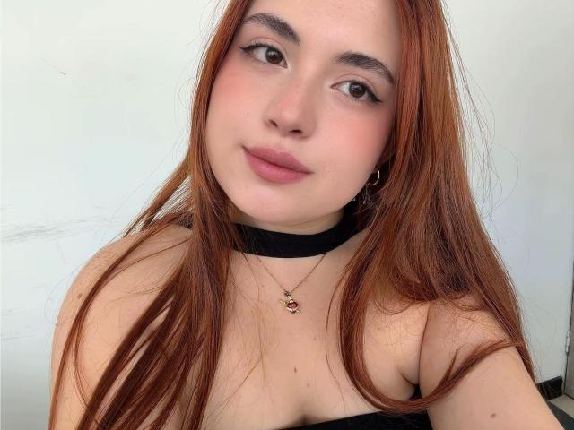 VicluX hat langes, rotes Haar, eine schwarze Halskette, dezentes Make-up, rosafarbene Lippen, kleine Ohrringe im Stecker-Stil und eine Halskette mit einem roten Stein. Ihre Brüste sind sichtbar, und sie schaut in die Kamera..