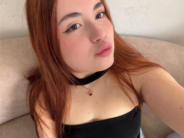 VicluX avec des lèvres pulpeuses, des cheveux bruns, un haut noir à bretelles, un collier noir et un pendentif rouge, regardant la caméra, peau claire, maquillage discret, eye-liner, rouge à lèvres rose clair, canapé beige..