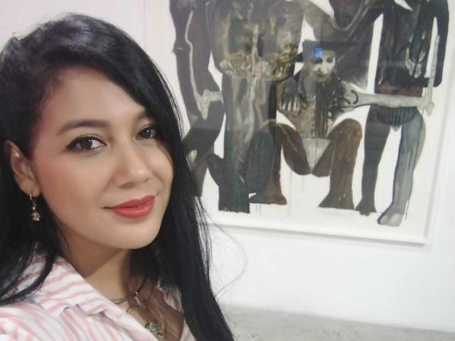 SophieEtty, com cabelos longos e pretos, batom vermelho e camisa listrada rosa, sorrindo para a câmera, usando pequenos brincos e um colar, em frente a uma pintura abstrata escura..