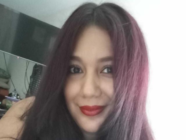 SophieEtty, avec ses cheveux teints en violet, son rouge à lèvres rouge et son sourire charmant, regarde légèrement sur le côté dans un autoportrait. Elle a un joli visage et de petites seins..