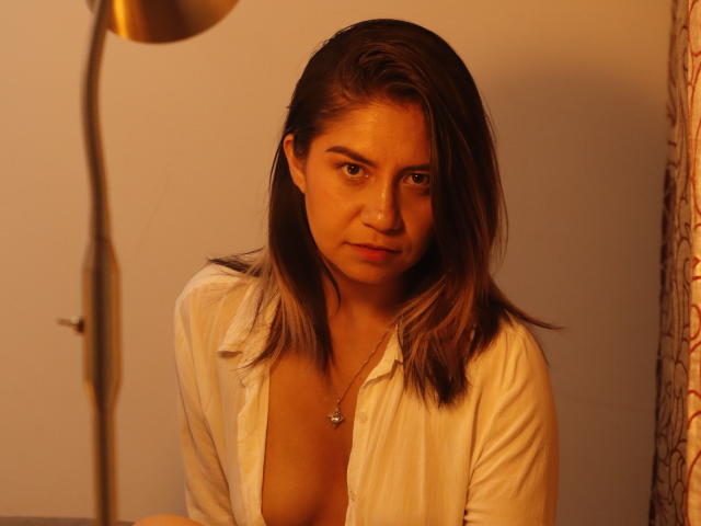 ValeriaLoren, con i capelli castani, una camicia bianca aperta, piccoli seni e un'espressione seria, seduta sotto una luce calda, indossa una collana con una piccola croce..