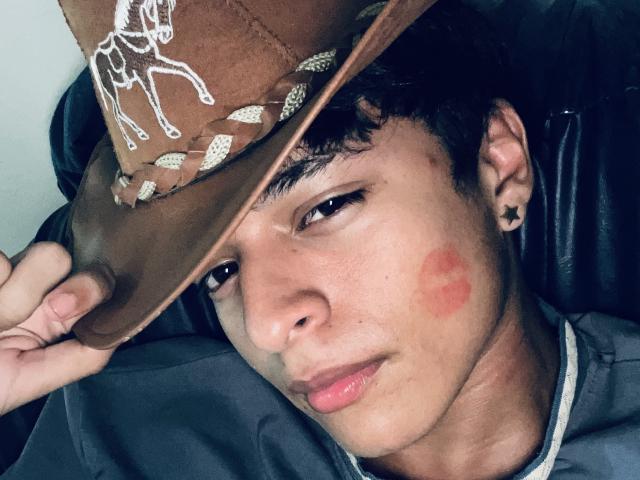 AbCampins, con un cappello da cowboy marrone, che lo tiene basso sugli occhi, ha un segno di bacio rosso sulla guancia destra, un orecchino a forma di stella nera e capelli corti e scuri, indossa una camicia grigia..