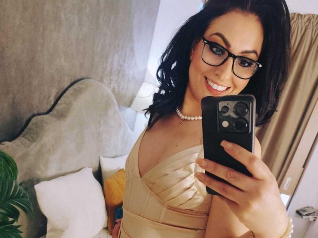 LisaCroftt, met zwarte bril, lachend, met een telefoon in haar hand, draagt een beige jurk met fronsjes, een parelketting en golvend zwart haar, en laat op een selfie haar borsten zien..