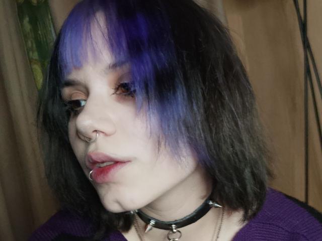 VickeyParkes has purple and black hair, a nose ring, lip ring, and a spiked choker. She's wearing a purple top with pale skin and a slightly open mouth.
