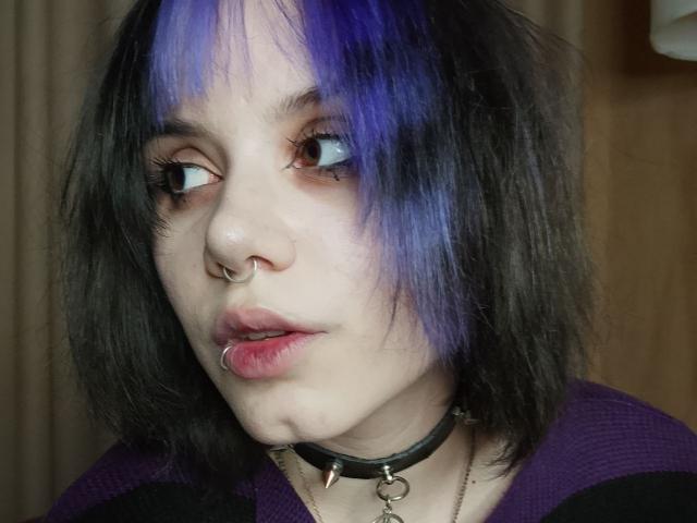 VickeyParkes with black and purple hair, nose ring, lip ring, and black choker with heart. Pale skin, looking to the side with slightly parted pink lips.