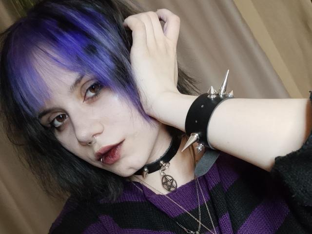 VickyParkes with purple and black hair, pale skin, black eye makeup, nose ring, lip piercing, black and purple striped shirt, spiked wristbands, and a pentagram necklace.