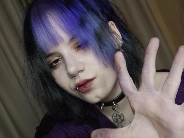 VickeyParkes with purple and black hair, nose ring, lip ring, choker with pentagram, and purple shirt, making a rock 'n' roll hand gesture, looking slightly off-camera.
