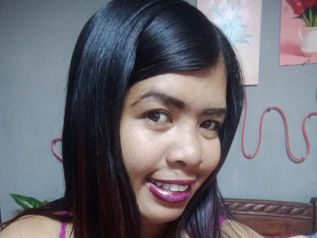MariahSquirting69 with shiny black hair, brown eyes, and purple lipstick, smiling at the camera. Her ass is not visible, but she's wearing a pink top.