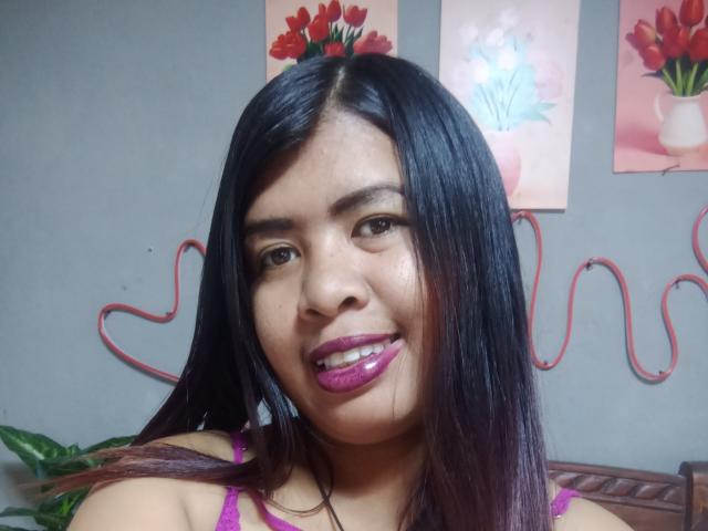 MariahSquirting69 with shiny black hair, purple lipstick, and purple bra, smiling at the camera, showing her cute face and small nose. Her tits are slightly visible.