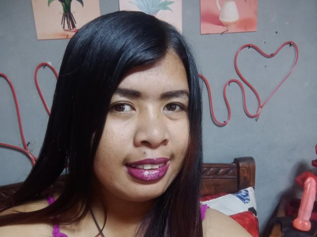 MariahSquirting69 has long black hair, brown eyes, and purple lipstick. She's wearing a pink top and has a nose piercing. A red heart-shaped rope is in the background.