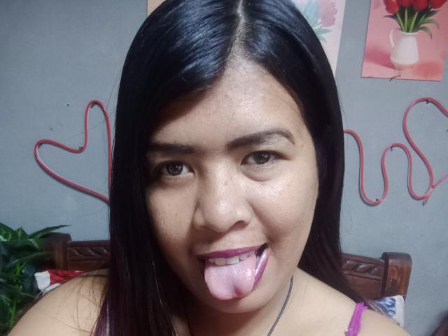 MariahSquirting69, with long black hair, brown eyes, and pink tongue sticking out, wearing a purple top, showing her face in a selfie.