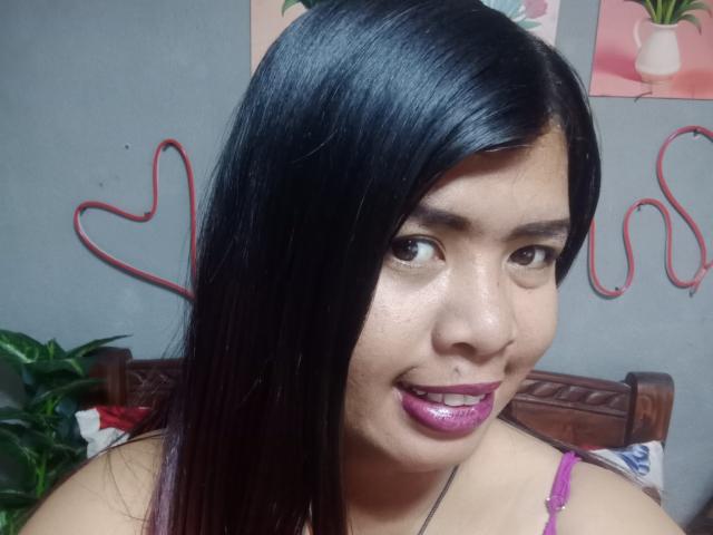 MariahSquirting69 with shiny black hair, purple lipstick, and a slight smile, showing her face and shoulders, wearing a pink strap, in a bedroom with heart decorations.