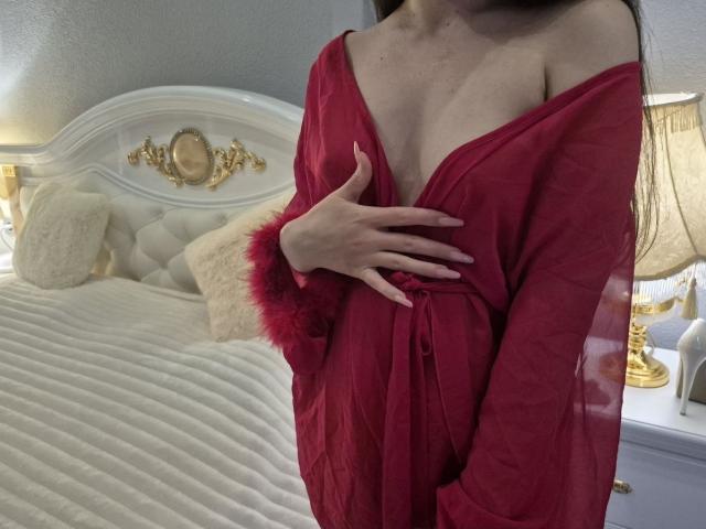 Rouzi porte une nuisette rouge, transparente, avec une épaule dénudée, et couvre ses seins d'une main, dans une chambre éclairée d'une lumière douce, avec un lit blanc capitonné et une lampe dorée..
