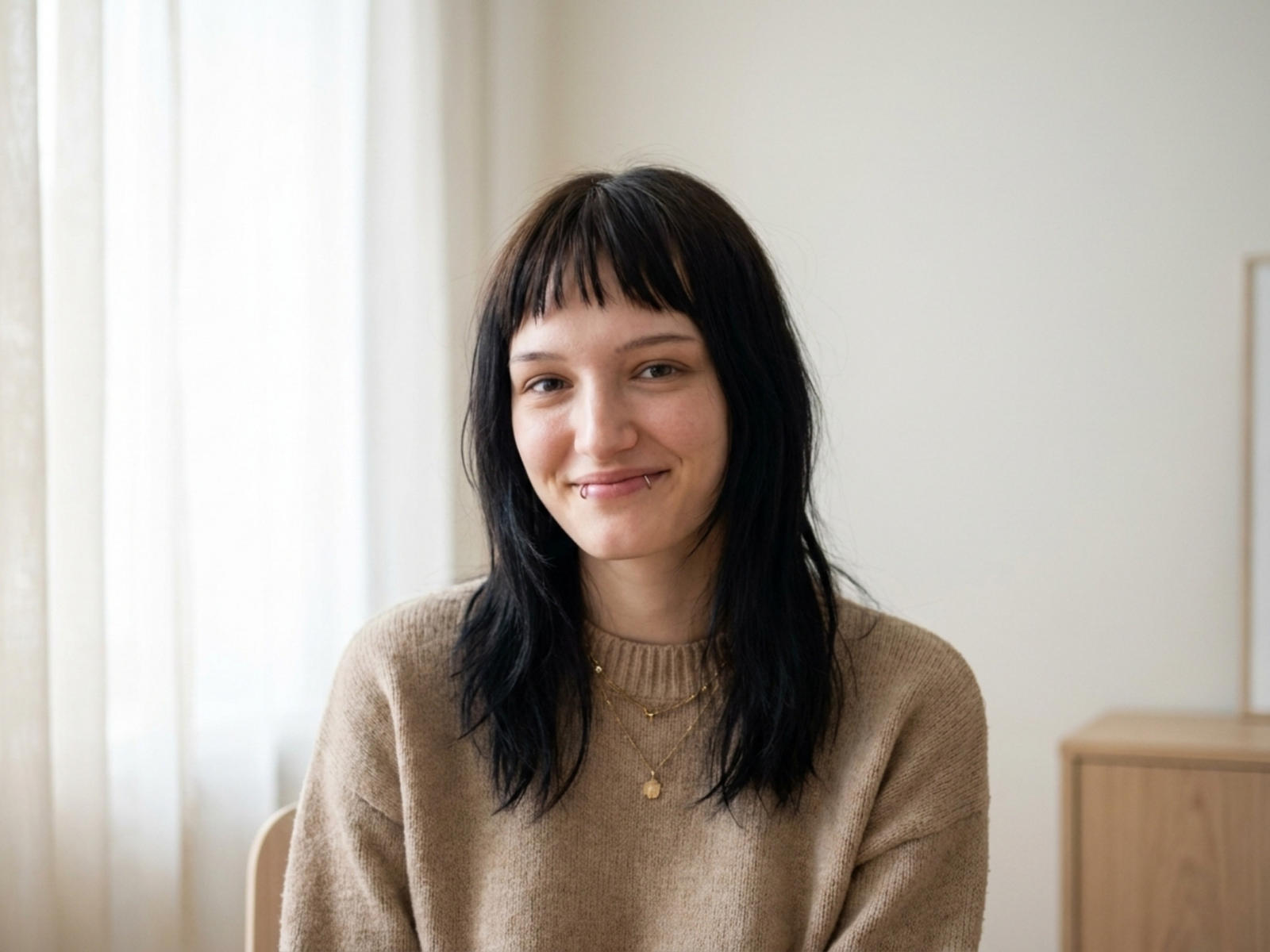 Valery Bazzle, con i capelli neri a metà schiena, sorridente, indossa un maglione beige, una piccola collana d'oro, è seduta, con una luce naturale soffusa, ha la pelle chiara, un aspetto casual, carino, rilassato, con i capelli leggermente spettinati..