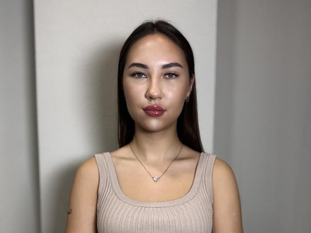 Alica Talhelm, avec de longs cheveux bruns foncés, portant un débardeur beige côtelé, regardant l'appareil photo avec des lèvres roses brillantes, un petit collier en forme de cœur et un maquillage discret..
