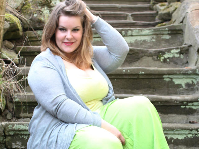 Una ragazza di nome Leny, con i capelli castano corti, indossa un cardigan grigio, una maglietta gialla e una gonna verde, seduta su gradini di pietra ricoperti di muschio, con una mano tra i capelli, sorridente, formosa, carina, con i glutei leggermente visibili..