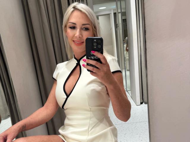 Cindy-Stone1-sensuelle, avec des cheveux blonds, une robe blanche avec des détails noirs et des ongles roses, se prend en photo dans un miroir, montrant sa poitrine, souriante, tenant un téléphone noir..
