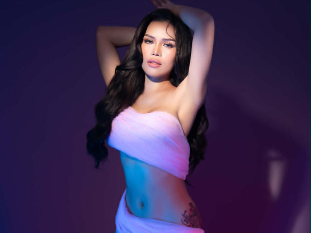 NikkiLuxe with long black hair, arms raised, wearing a pink strapless towel covering her tits, white towel wrapped around her ass, purple-blue lighting, tattoo on her right hip.