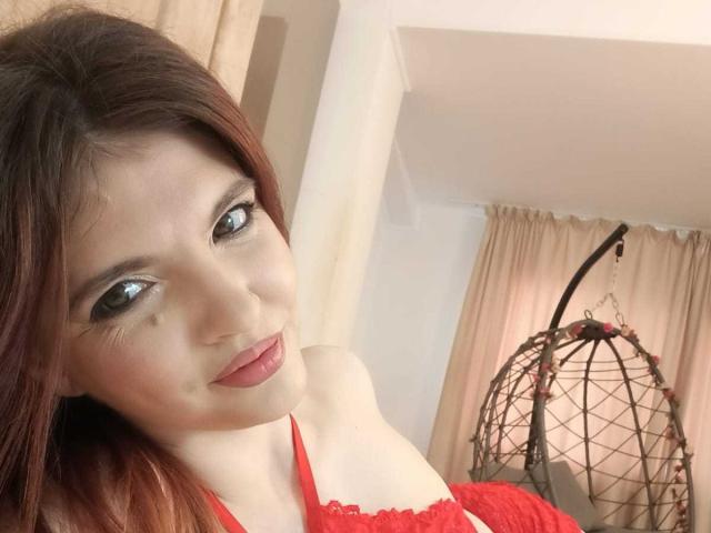 SinfulTina con lápiz labial rojo, ojos verdes y una blusa de encaje rojo, mostrando su hombro descubierto y un ligero escote, mirando a la cámara con una sonrisa sutil..
