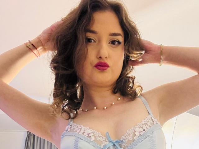 JessyAmour with curly brown hair, pink lipstick, wearing a blue lace bra with white trim, hands in hair, gold bracelet, pearl necklace, and pink bracelet, looking at camera.