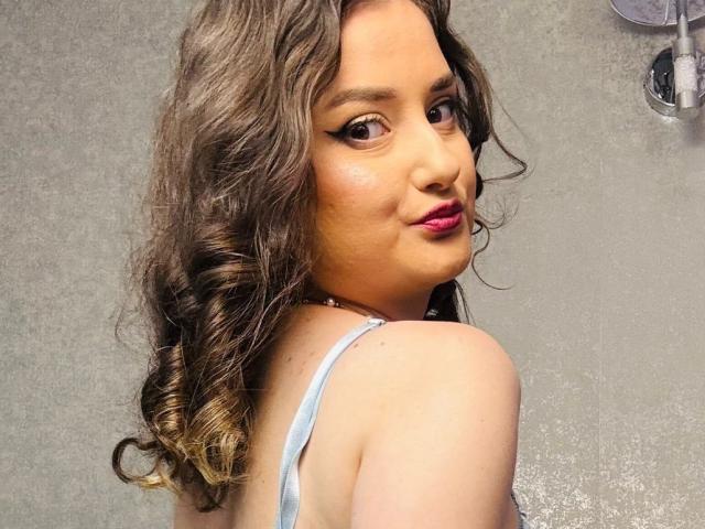 JessyAmour with curly brown hair, wearing a light blue top, showing her back and looking over her shoulder with pink lipstick. Her tits are partially visible.