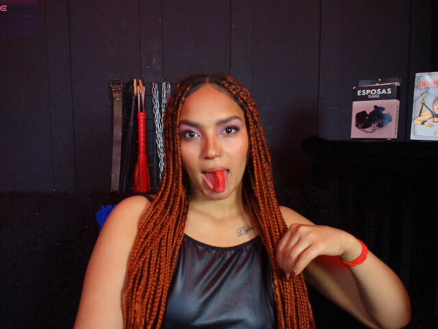 BrianaPratt with long orange braids, black leather top, tongue out, and red wristband, holding braid, looking at camera. Visible tattoo on chest. BDSM whip and bookshelf in background.