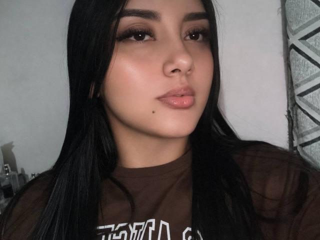 AmaliaDalton has long black hair, brown eyes, full pink lips, a small beauty mark on her right cheek, and wears a brown shirt. Her nose is pierced.