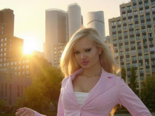 Selena Altringer, con i capelli biondi, un blazer rosa e una camicetta bianca, che mette in mostra il suo seno, in piedi di fronte ai grattacieli della città al tramonto, appare sexy e sicura di sé..