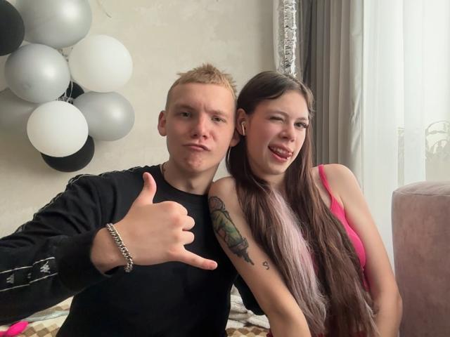MorningRubbing met blond haar, zwart shirt en armband, die een duimpje omhoog steekt, naast MorningRubbing met bruin haar, een roze top en een getatoeëerde arm, die lacht. Ballonnen op de achtergrond..