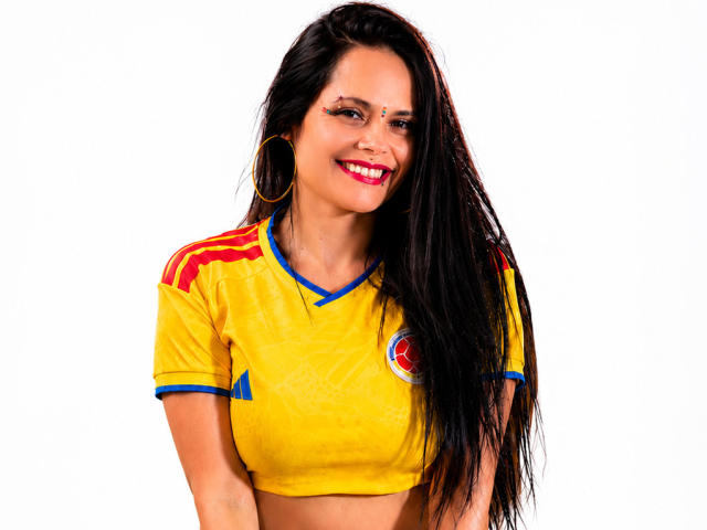 CharlotteCrofort with long black hair, yellow sports shirt, red and blue accents, gold hoop earrings, smiling, showing red lipstick, medium-sized tits, white background.