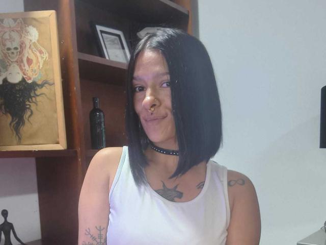 Amelie Blake, com cabelo curto preto, piercing no nariz, colar, top branco, tatuagens no pescoço e no braço, sorrindo, pele morena, em um quarto com prateleiras de madeira e obras de arte..