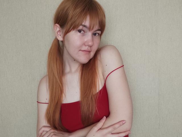 SheritaChieng, avec des couettes rousses, portant un haut rouge sans bretelles, les bras croisés, un léger sourire, une peau pâle, de petites seins et des yeux marrons, sur un fond beige uni..