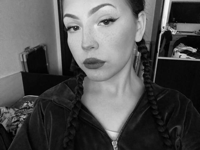 Nadine Waddick tem pele com sardas, cabelo escuro trançado, sobrancelhas grossas, delineador nos olhos e lábios cheios. Ela está vestindo uma blusa escura com zíper e está olhando para a câmera com uma expressão séria..