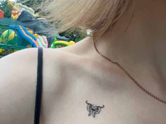 Tora Stambaughs freie Schulter mit einem schwarzen Schmetterling-Tattoo, einem schwarzen Spaghetti-Träger-Top und einer goldenen Kette, wobei ihr hellbraunes Haar zu sehen ist..