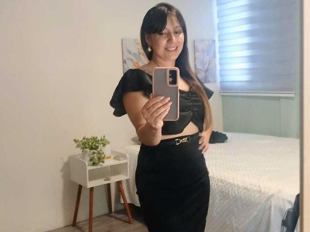 RoxanneBlair with long brown hair, black off-shoulder top, black skirt, holding phone, smiling, standing in bedroom, mirror selfie, visible boobs, small gold belt, white bed, plant on side.