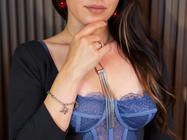 AureliaNoir mit der Hand in der Nähe ihres Kinns, trägt einen blauen Spitzen-BH, ein schwarzes Oberteil, rote Ohrringe, eine silberne Halskette und ein Armband, wobei ihre Brüste sichtbar sind und sie einen Ring mit einem Kreuz trägt..