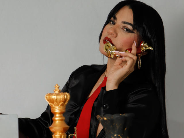 AliceCarusso with long black hair, red lipstick, and a gold phone, wearing a black satin jacket and red tie, holding a gold crown and a black figurine.