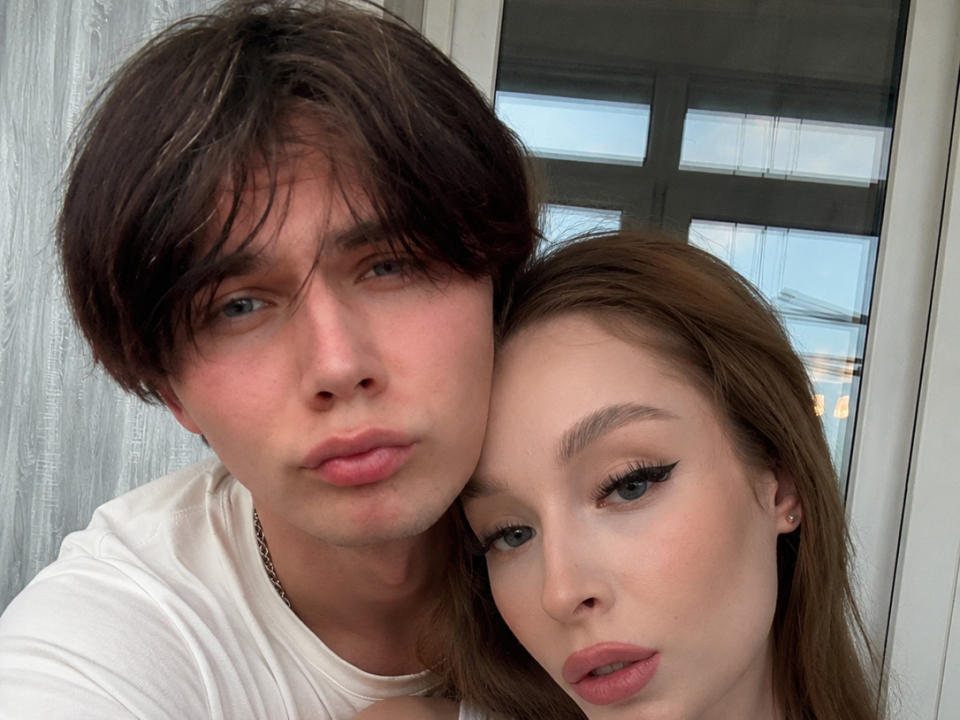 SherlyDewick, con cabello castaño corto y ojos azules, y SherlyDewick, con cabello castaño largo y delineador de ojos, ambas haciendo pucheros, en una camisa blanca, en un selfie..
