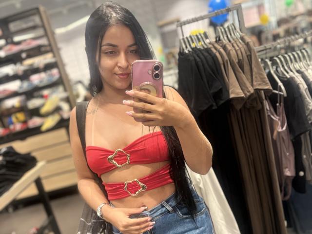 HikariChloe with long black hair, red strapless top with gold rings, showing her tits, holding a pink phone, standing in a store, wearing blue jeans.