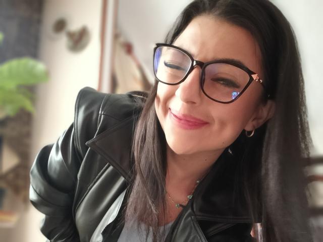 Anntonella Watson, met lang zwart haar, zwarte bril en een zwarte leren jas, lacht met roze lippenstift, en laat een schattig gezicht en een kleine neus zien, terwijl ze oorringen draagt..