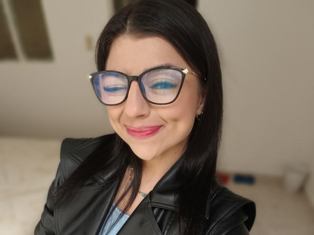 Anntonella Watson met zwarte bril, blauwe eyeliner, roze lippenstift, een zwarte leren jas, die lacht, met recht zwart haar, een lichte huid, kleine oorringen en een subtiele tatoeage op haar nek..