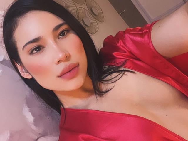 TamaraSsummer heeft een gladde, lichte huid, zwart haar en draagt een rode satijnen jurk met een diepe halslijn, waardoor haar decolleté zichtbaar is. Ze heeft roze blush en subtiele make-up..