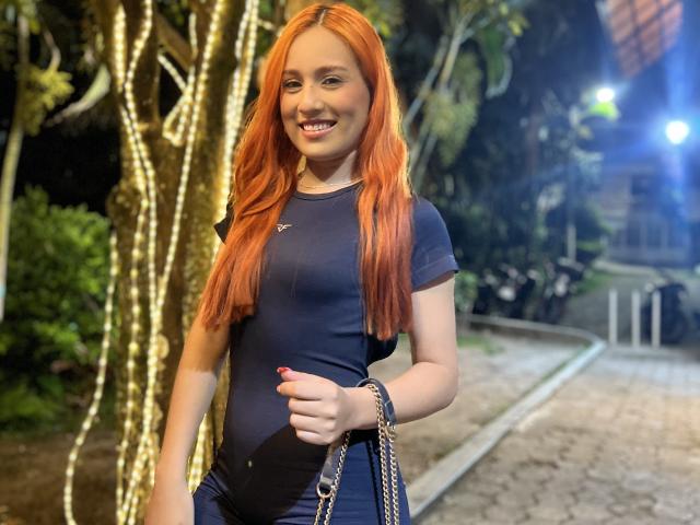 EmmaWat avec des cheveux roux, un t-shirt bleu moulant, de petites seins, un ventre de grossesse, souriante, tenant un sac à main, debout à l'extérieur, nuit, arbre, lumière en arrière-plan, décontractée, attirante, mignonne..