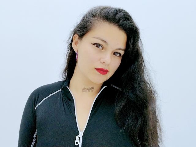Ihris tem cabelos pretos longos, batom vermelho e veste uma blusa preta com zíper e detalhes brancos, mostrando uma pequena tatuagem no pescoço. Seus seios não estão visíveis..
