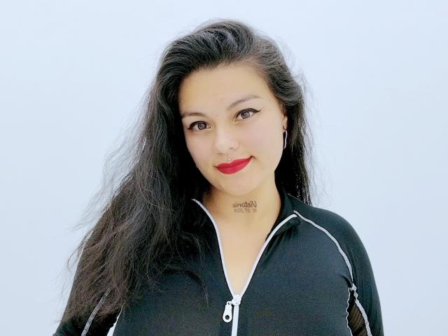 Ihris ha lunghi capelli neri, rossetto rosso e indossa una giacca nera con zip e strisce bianche. Sorride alla fotocamera e ha un tatuaggio sul collo..