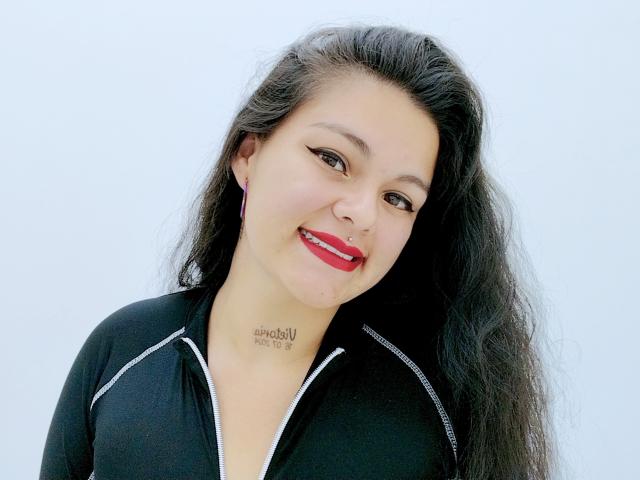 Ihris hat langes, schwarzes Haar, roten Lippenstift und ein schwarzes, vorne zugezipptes Hemd. Sie lächelt und hat ein Tattoo am Hals. Sie trägt rosa Ohrringe. Ihre Brüste sind klein..