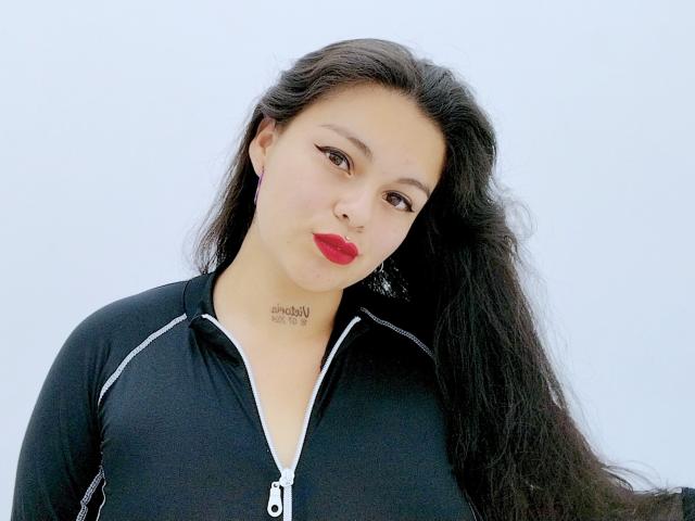 Ihris hat langes, schwarzes Haar, roten Lippenstift und trägt ein schwarzes Top mit Reißverschluss. Ihre Brüste sind klein, und sie hat ein Tattoo im Nacken. Sie schaut in die Kamera..