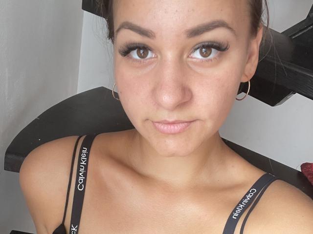 RoseStrawberriies tem olhos castanhos, pele clara e pequenas argolas nos ouvidos. Ela está usando um sutiã preto da Calvin Klein, com os seios levemente visíveis. Ela está olhando para a câmera com um sorriso discreto..