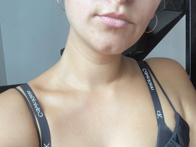 RoseStrawberriies hat helle Haut, rosafarbene Lippen, kleine Ohrringe im Stecker-Stil und trägt einen schwarzen Calvin Klein-BH mit sichtbaren Trägern. Ihr Schlüsselbein ist leicht hervorgehoben..